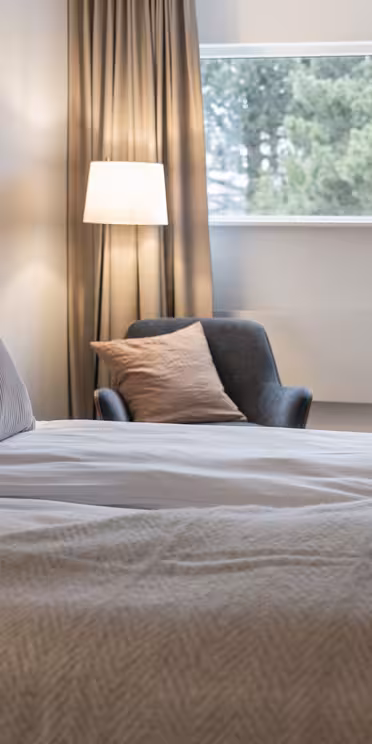 Hotellrom med stor seng, hvitt og beige sengetøy, og en svart lenestol ved en høy lampe. Et trepult med stol og et vindu med utsikt over trærne fullfører den koselige, nøytrale toneplassen.